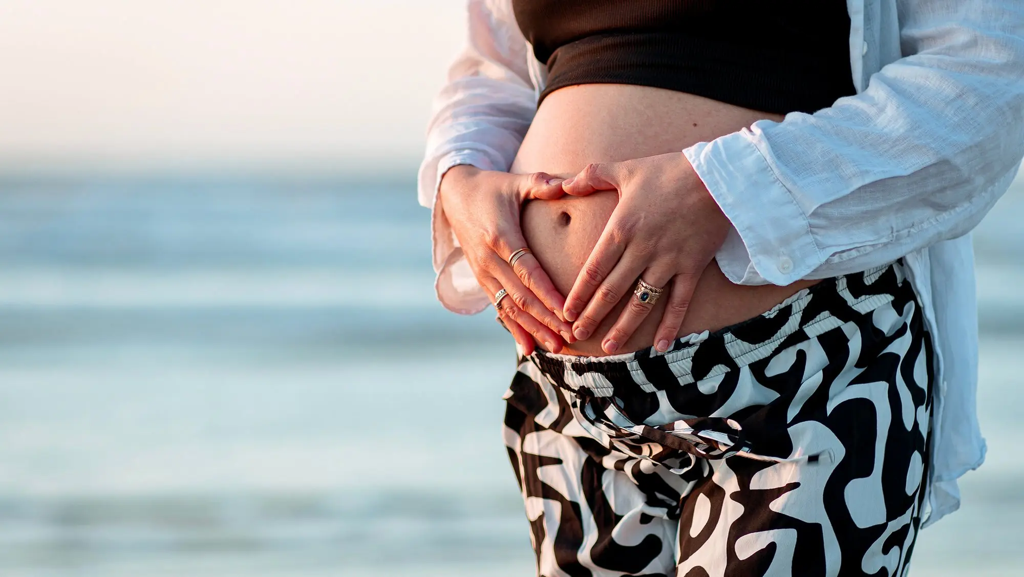 Hands making a heart shape over a pregnant bump, with the sea behind.