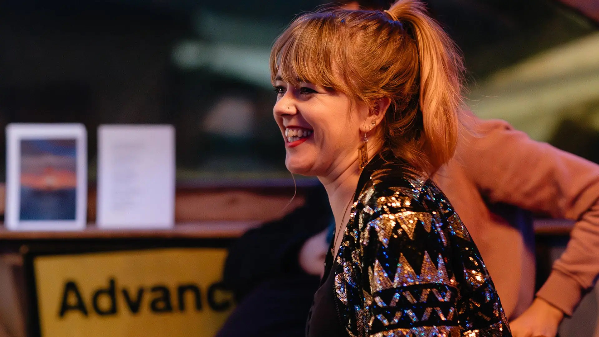A woman in a sequinned jacket laughs at a table during an evening of live music.