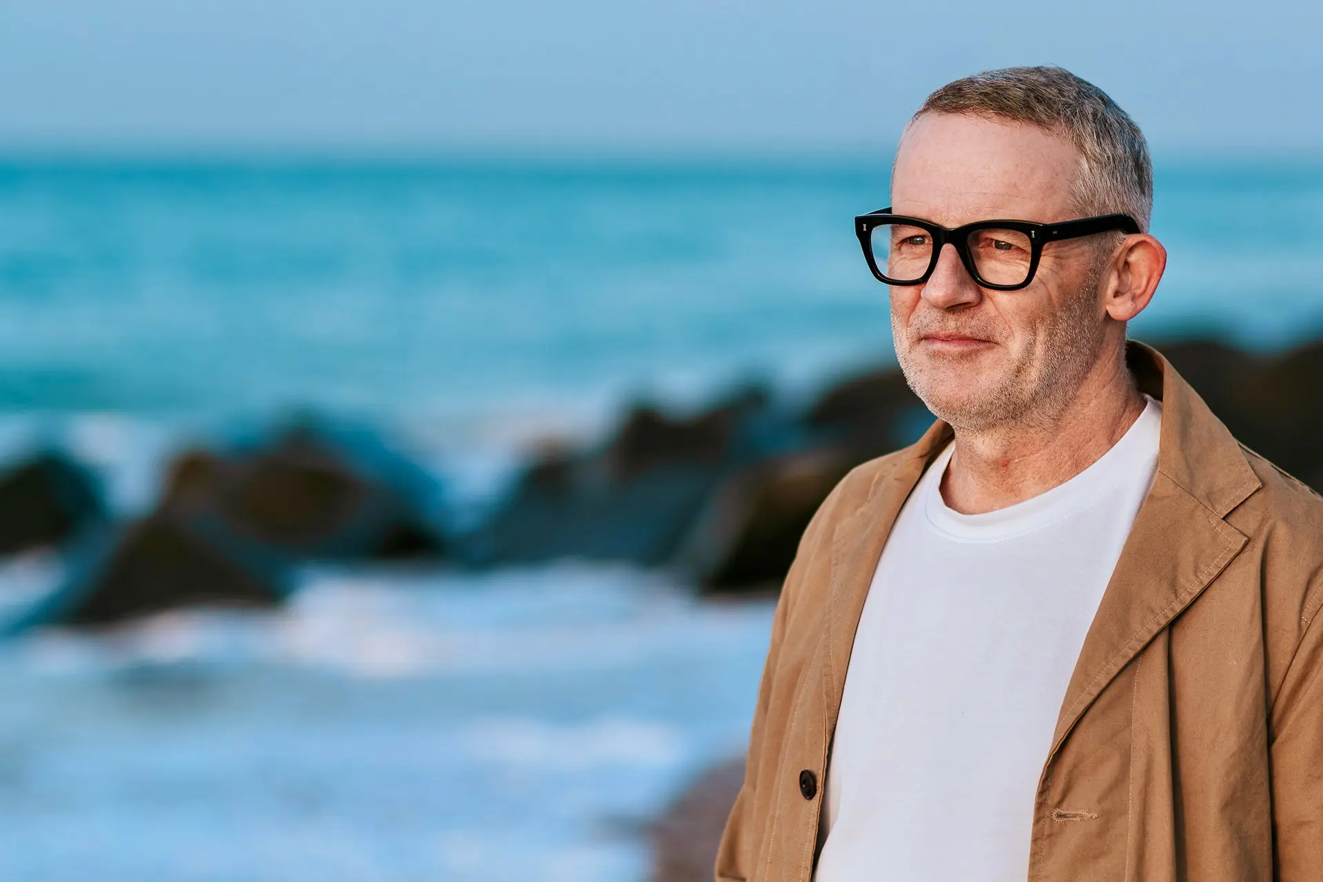 Man in a tan jacket smiles gently on the shingle beside a turquoise sea.