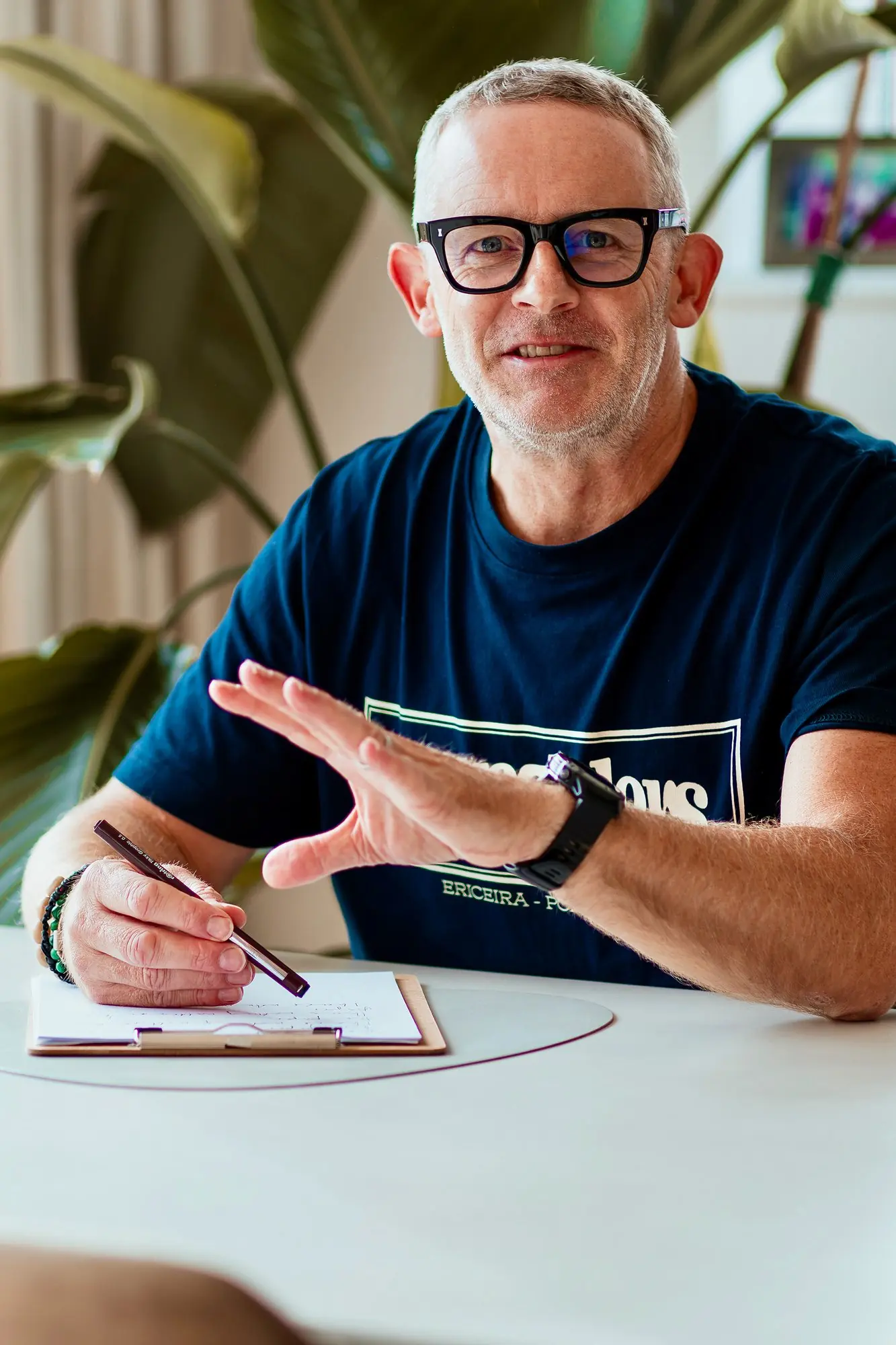 Man in a navy t-shirt writes at a desk beside a large green houseplant.