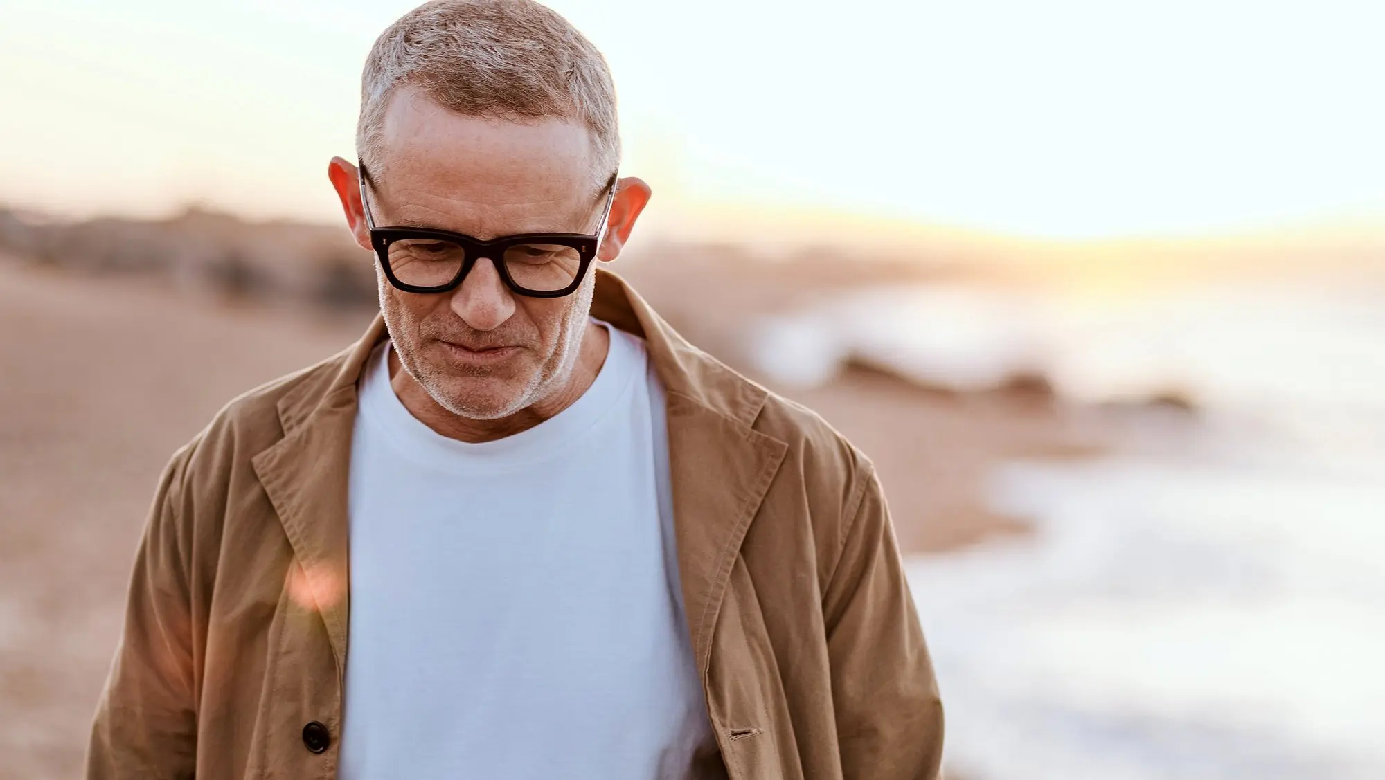 A man in glasses and a tan jacket looks down on a beach at sunrise.