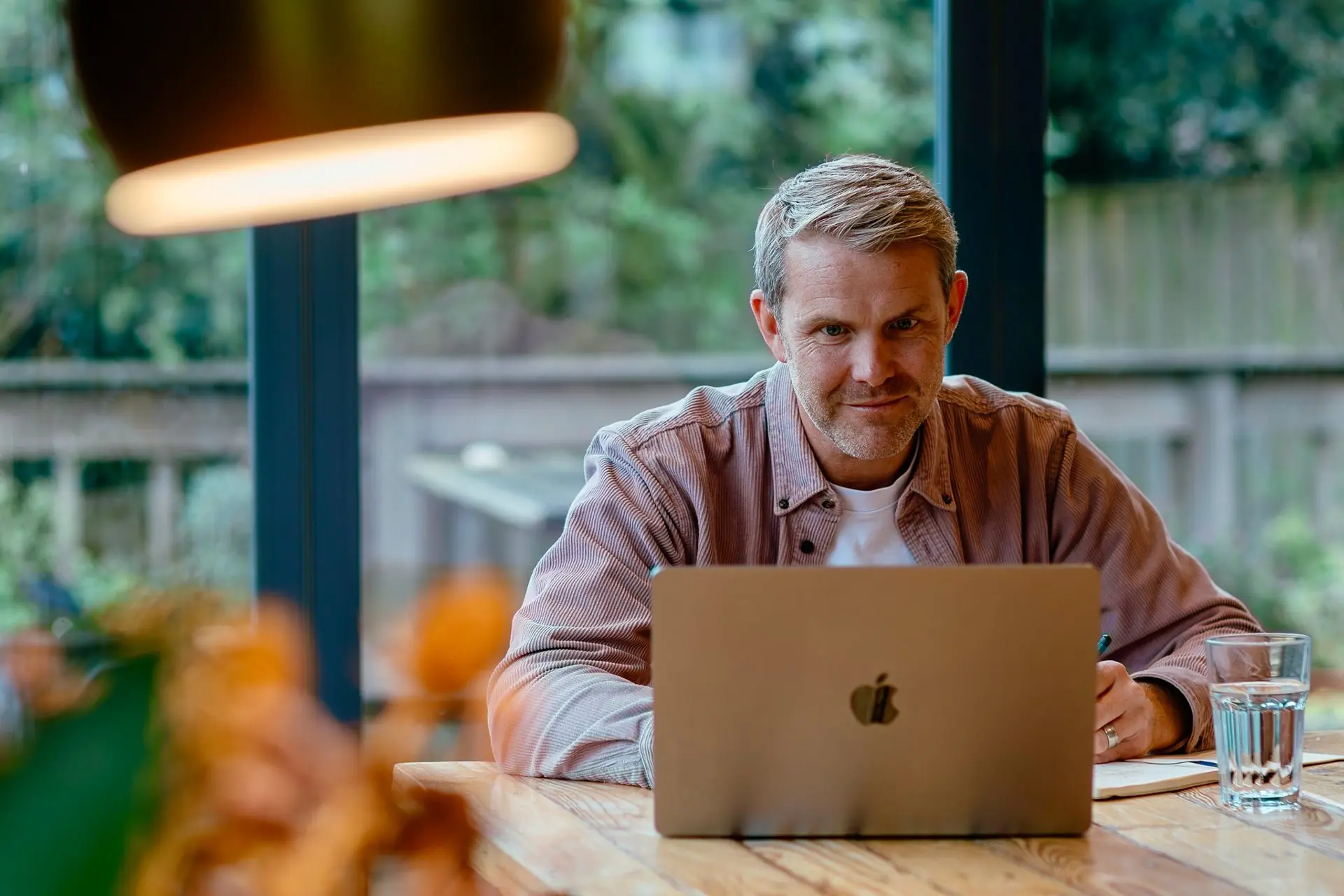 Man sits at a wooden table with a laptop and a glass of water.