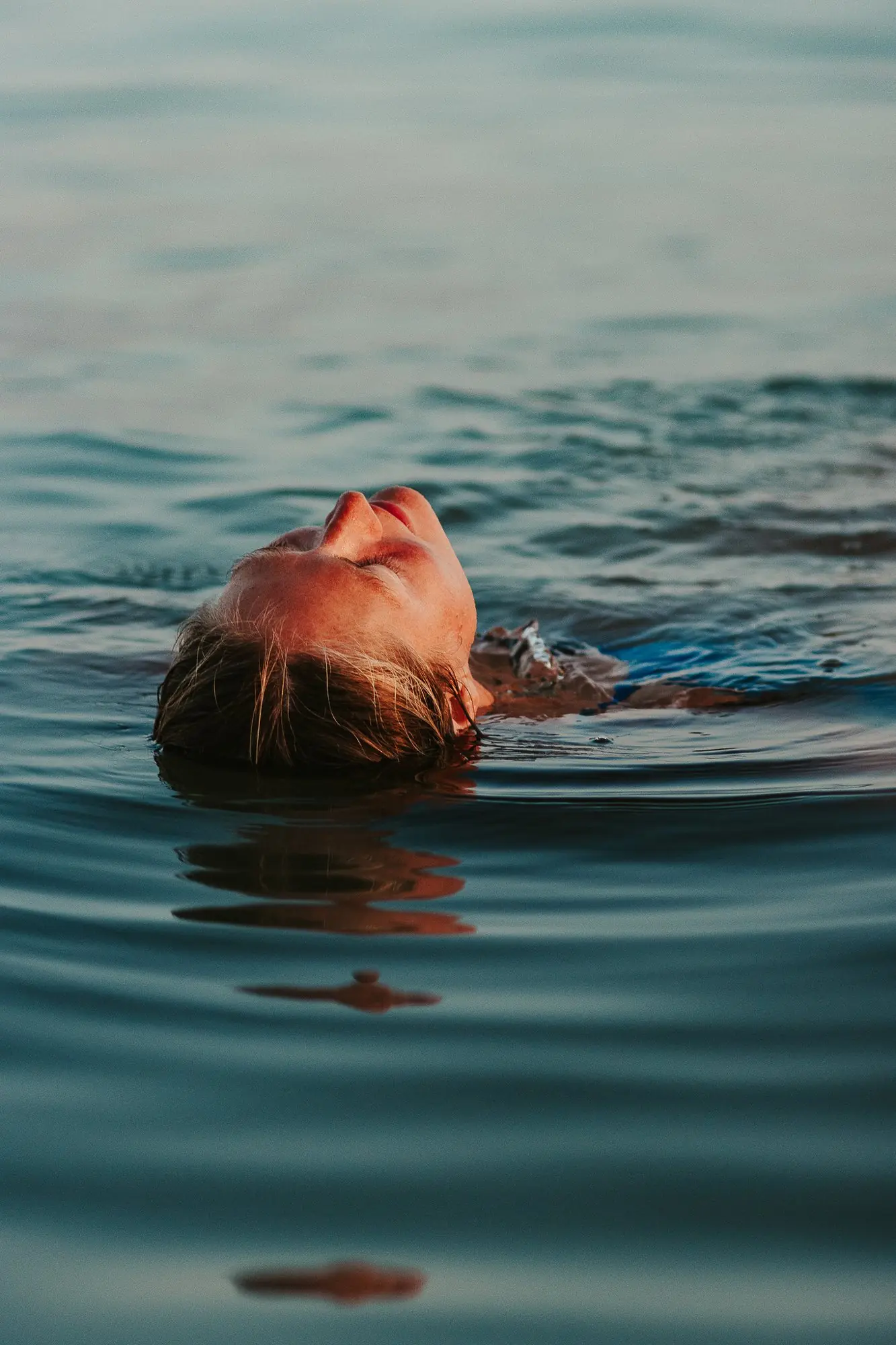 A woman floats on her back in calm sea at sunset, eyes closed, her face lit by warm evening light.