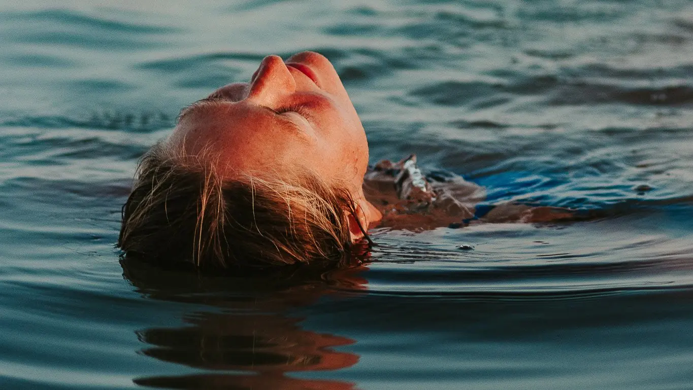 A woman floats on her back in calm sea water, eyes closed.