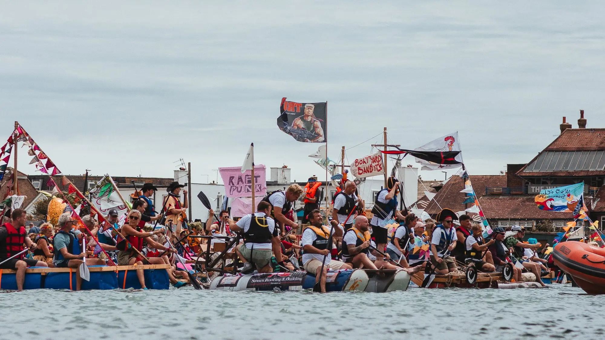 Crowded homemade rafts with flags and bunting on the river at Shoreham.