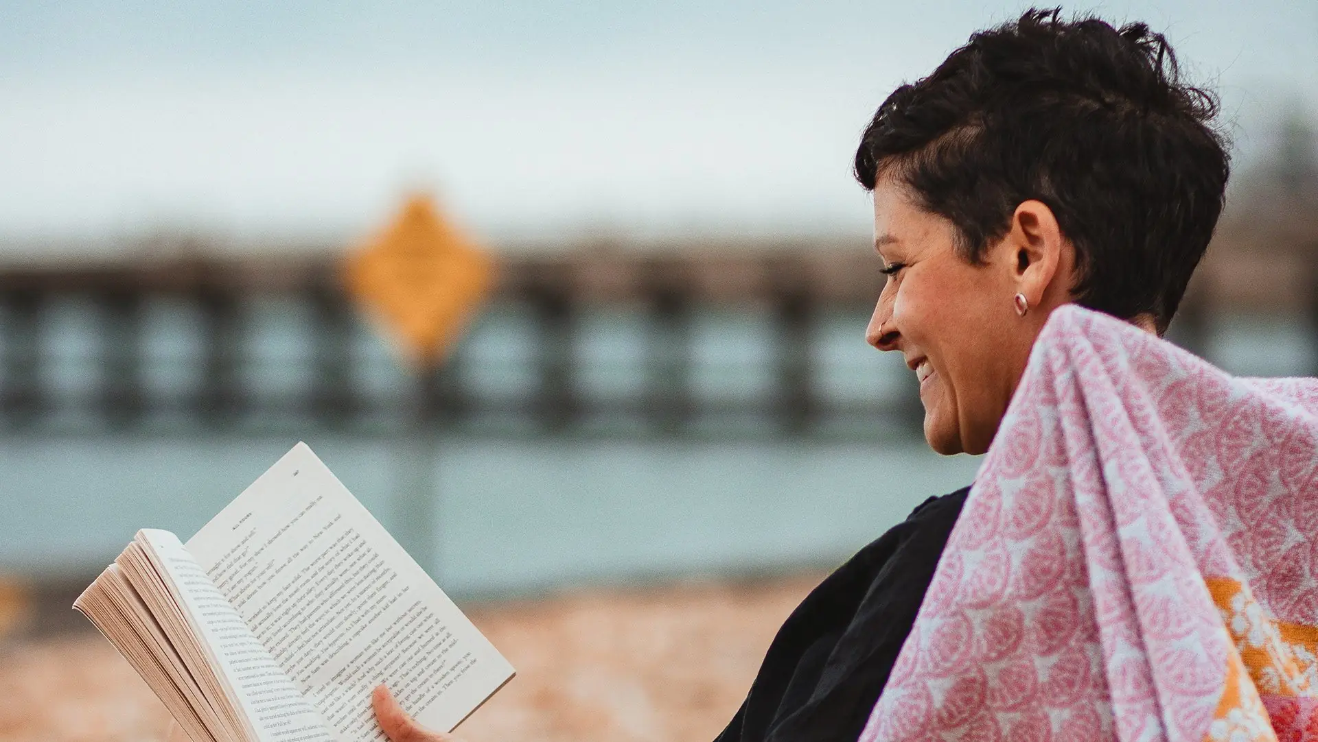 A smiling woman wrapped in a pink towel reads a book by the water.