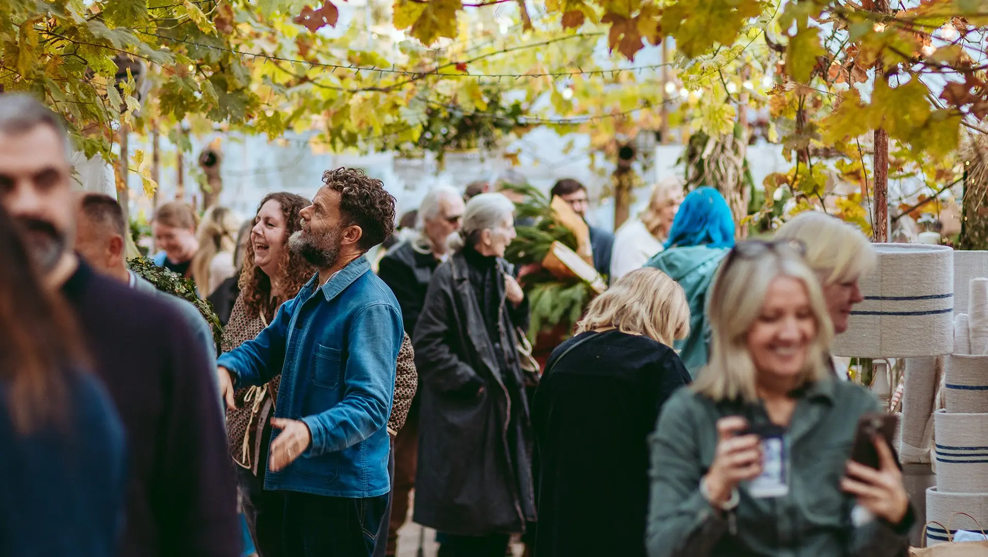 A crowd of visitors chatting and smiling under autumn vines at a busy fair.